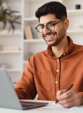 A medium-skinned man in an orange shirt looks at a laptop on a desk.