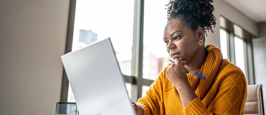 A black woman in a mustard yellow sweater sits looking at a laptop.